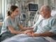 A young Caucasian female respiratory therapist in light scrubs sits beside an anxious male ICU patient, offering reassurance in a modern hospital room with monitors and equipment in the background.